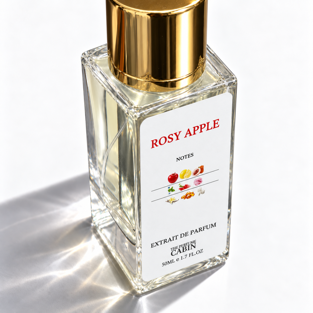 A 50ml glass bottle of 'Rosy Apple' extrait de parfum by 'The Perfume Cabin', featuring a white label with red text and fruit illustrations, topped with a shiny gold cap, set against a bright white background with soft shadows.