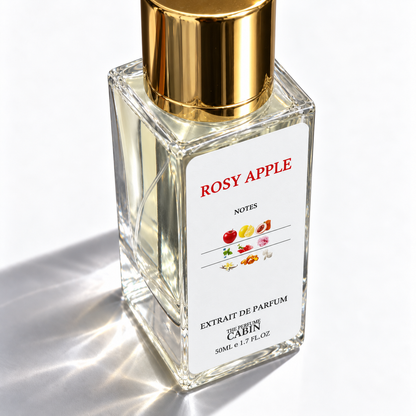 A 50ml glass bottle of 'Rosy Apple' extrait de parfum by 'The Perfume Cabin', featuring a white label with red text and fruit illustrations, topped with a shiny gold cap, set against a bright white background with soft shadows.