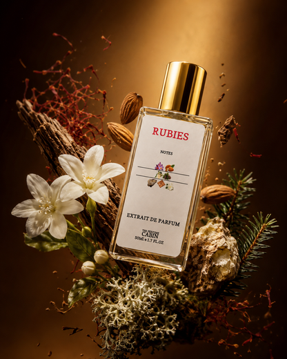 A bottle of RUBIES Extrait de Parfum, Noüreau, with a gold cap and white label, surrounded by white flowers, pine needles, and spices on a dark brown background.
